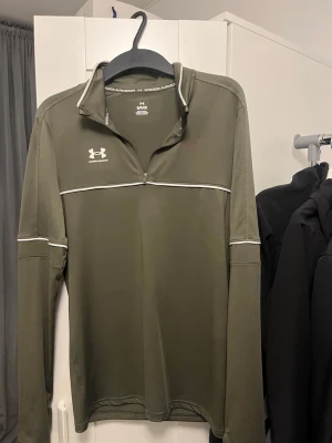 Olivgrön träningströja Under Armour - Snygg olivgrön långärmad träningströja från Under Armour med halv dragkedja och vita detaljer. Tillverkad i ett lätt och snabbtorkande funktionsmaterial som passar perfekt till löpning eller gym. Logga på bröstet och ryggen.