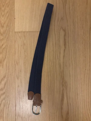 Mörkblått flätat bälte med skinn - Snyggt mörkblått flätat bälte med detaljer i brunt skinn och ett silvrigt spänne. Perfekt accessoar för att lyfta din outfit och funkar till både jeans och chinos. Enkel att justera och ger en stilren look.