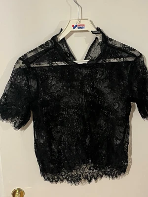 Svart spetsblus med kort ärm - Säljer en svart blus i genombruten spets med korta ärmar och hög hals. Blusen har fina spetsdetaljer och är lätt transparent, perfekt för att lyfta vilken outfit som helst. Pris kan diskuteras vid snabb affär