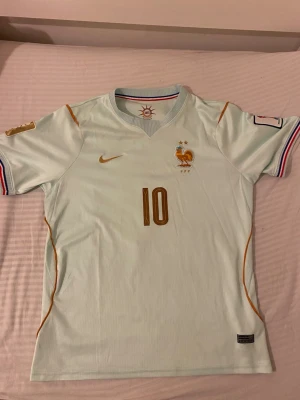 Frankrike fotbollströja  - Snygg ljusblå fotbollströja från Nike med Frankrikes landslagsemblem och nummer 10 i guld på bröstet. Tröjan har gulddetaljer, korta ärmar med randiga muddar och broderade märken. Perfekt för match eller träning.