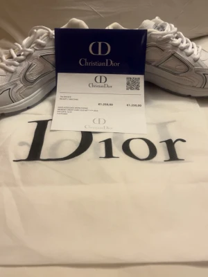 Helt Äkta Vita Dior sneakers med silverdetaljer Reflexfri - Snygga vita Dior sneakers med stilrena silverdetaljer och tydlig Dior-logga på sidan. Skorna har en chunky sula och är tillverkade i en mix av läder och textil. Perfekta för dig som vill sticka ut med exklusiva och trendiga sneakers.