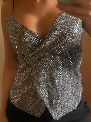 Glittrig silver omlottopp - Säljer en glittrig silverfärgad omlottopp med tunna axelband och djup v-ringning. Toppen har ett glittrande mönster och är perfekt för dig som vill sticka ut. Materialet känns lätt och följsamt mot kroppen.