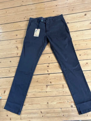 Mörkbruna chinos i slim fit - Snygga mörkbruna chinos i slim fit-modell med klassiska fickor och bälteshällor. Byxorna har en stilren design och är tillverkade i bomull med en liten del elastan för extra komfort. Perfekta för en trendig och avslappnad look.