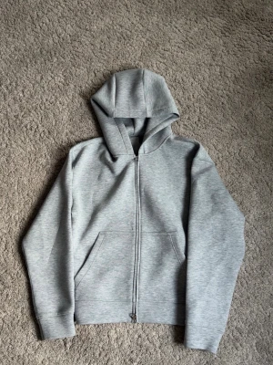 Kvalité Ljusgrå zip hoodie. - Säljer en stilren ljusgrå hoodie med dragkedja framtill och stor ficka på magen. Hoodien har en klassisk huva och ribbade muddar vid ärmsluten. Perfekt för en avslappnad och enkel look. Materialet känns mjukt och bekvämt. Och är jättebra kvalité. Storlek M.