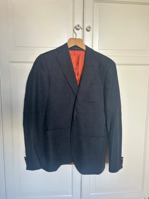 Mörkgrå Morris kavaj med orange foder - Stilren mörkgrå kavaj med snyggt orange sidenfoder. Klassisk passform med två framfickor och breda slag. Perfekt för dig som vill ha en tidlös och elegant look. Kavajen har dekorativa knappar vid ärmsluten och en innerficka med randig kant.