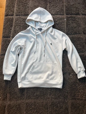 Vit hoodie från Polo Ralph Lauren - Snygg vit hoodie från Polo Ralph Lauren i storlek S. Klassisk design med huva, dragsnören och känguruficka framtill. Liten broderad logga på bröstet. Tillverkad i mjukt material för en skön och avslappnad look.