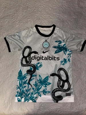 Inter Milan matchtröja med ormmotiv - Unik Inter Milan matchtröja från Nike i vitt och svart med turkosa detaljer. Tröjan har korta ärmar, svart krage och muddar samt ett iögonfallande ormmotiv och bladmönster på framsidan. Digitalbits-logga på bröstet och klubbmärke i turkost. Perfekt för fotbollsfans!