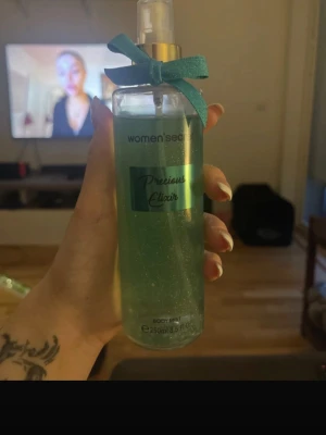 Precious Elixir Body Mist - Fräsch body mist från Women'secret i doften Precious Elixir. Flaskan är genomskinlig med turkost innehåll och glittereffekt, dekorerad med en söt turkos rosett vid spraymunstycket. Rymmer 250 ml och har en elegant, smal form som är enkel att hålla i.