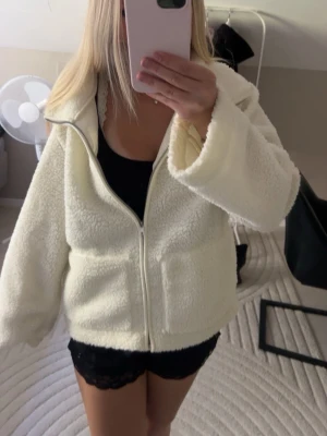 Vit teddyjacka med dragkedja - Mysig vit teddyjacka med dragkedja framtill och stor krage. Jackan har en oversized passform och är gjord i fluffigt, mjukt material som ger en riktigt cozy känsla. Perfekt för kyliga dagar när du vill vara både varm och stylish. Storleken är M men skulle säga att det passar som en S också💓💓💓
