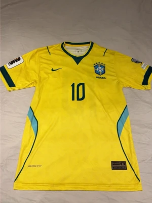 Gul Brasil Neymar fotbollströja - Säljer en gul Brasilien fotbollströja från Nike med Neymar Jr. och nummer 10 på ryggen. Tröjan har blå detaljer, officiell landslagssköld på bröstet och Aero-Fit material för extra komfort. Perfekt för dig som älskar fotboll och vill stötta Brasilien.
