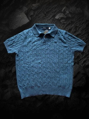 Ralph Lauren stickad piké tröja  - Säljer en blå stickad skjorta från Polo Ralph Lauren med korta ärmar och klassisk krage. Skjortan har ett diskret kabelstickat mönster och knappar framtill. Perfekt för dig som vill ha en stilren och bekväm look. Har aldrig använts 