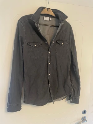 Mörkgrå skjorta från ASOS DESIGN - Snygg mörkgrå slim fit skjorta från ASOS DESIGN i bomull. Skjortan har långa ärmar, klassisk krage och tryckknappar framtill. Två bröstfickor med lock och knappdetaljer ger en cool touch.