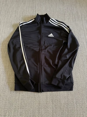 Svart Adidas träningsjacka - Säljer en svart träningsjacka från Adidas med dragkedja framtill och klassiska vita ränder längs ärmarna. Jackan har ribbade muddar och Adidas-logga på bröstet. Perfekt för träning eller chill. 