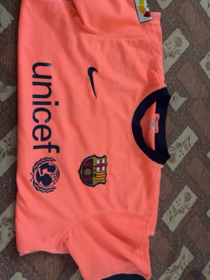 Barça orange matchtröja Nike - Säljer en orange FC Barcelona matchtröja från Nike med mörkblå detaljer. Tröjan har klubbmärke, Nike-logga och UNICEF-tryck på framsidan. Rund halsringning med kontrastfärg och korta ärmar. Perfekt för fotbollsträning eller som supporterplagg.
