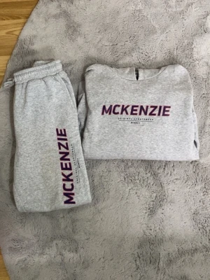 Grått loungewear-set från McKenzie (12-13år) - Mysigt grått loungewear-set från McKenzie med matchande sweatshirt och byxor. Båda plaggen har lila McKenzie-tryck framtill. Perfekt för chill dagar hemma. Mjukt material och avslappnad passform.