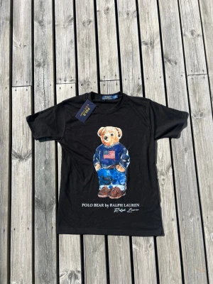 Svart Polo Bear t-shirt från Ralph Lauren - Svart t-shirt från Ralph Lauren med Polo Bear-tryck framtill. Klassisk passform med rund hals och korta ärmar. Tillverkad i mjuk bomull, perfekt för en avslappnad och cool stil.