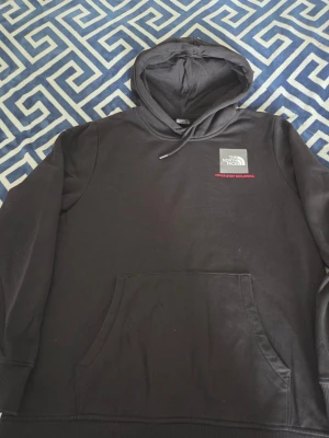 Svart hoodie från The North Face - Säljer en svart hoodie från The North Face med klassisk logga på bröstet och texten 'Never Stop Exploring' i rött. Hoodien har huva med dragsko och en stor magficka. Perfekt för chill dagar och streetwear vibes.