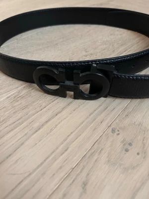 Svart skärp med mattsvart spänne - Snyggt svart skärp i skinn med ett stilrent mattsvart metallspänne i en unik design. Perfekt accessoar för att lyfta din outfit och ge en clean look. Skärpet har en slät yta och klassisk form.