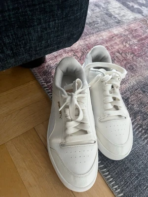 Vita sneakers från Puma - Stilrena vita sneakers från Puma med klassisk låg siluett. Skorna har vita snören, diskreta perforeringar på tån och subtila detaljer i vitt och grått. Perfekta för en clean och sportig look.