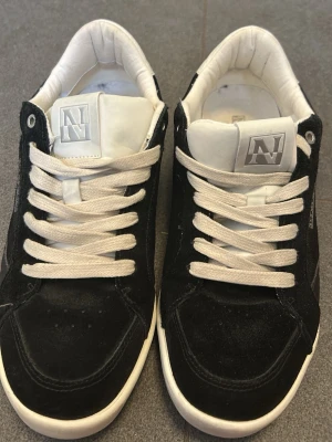 NapaPijri Sneakers - Snygga sneakers i svart mocka med vita detaljer och vita skosnören. Skorna har en vit logga på plösen och vit sula. Insidan är vadderad för extra komfort och designen är klassiskt låg. Perfekta för en stilren och avslappnad look.