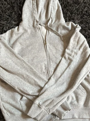 Ljusgrå zip-hoodie med huva - En stilren ljusgrå hoodie med dragkedja framtill och klassisk huva med dragsko. Tröjan har ribbade muddar vid ärmslut och nederkant samt två fickor framtill. Perfekt för en avslappnad och bekväm stil.