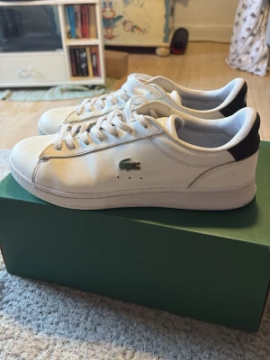 Vita Lacoste sneakers med svart detalj - Säljer ett par klassiska vita Lacoste sneakers med lågt skaft och svart detalj vid hälen. Skorna har den ikoniska gröna krokodilen på sidan och vita snören. Tillverkade i slätt läder med diskreta sömmar och en stilren look. Hör av vid frågor. Pris kan diskuteras!
