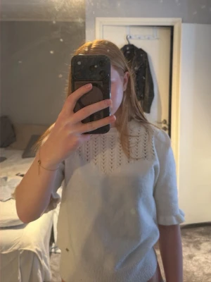 Vit stickad tröja med kort ärm - Säljer en söt vit stickad tröja med korta ärmar och ett hålmönster upptill vid halsen. Tröjan har en rund halsringning och ribbstickade muddar vid ärmslut och nederkant. Perfekt för lager-på-lager och passar till jeans eller kjol.