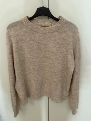 Beige/ljusrosa stickad tröja  - Beige ljusrosa färgad stickad tröja med rund hals och långa ärmar. Tröjan har ribbade muddar vid ärmslut och nederkant, vilket ger en snygg och avslappnad look. Perfekt att matcha med jeans eller kjol för en casual vibe.