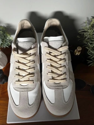 Maison Margiela Gats - Snygga sneakers i vitt och beige med ovandel i mocka och släta partier i läder. Skorna har beige snörning och en klassisk gummisula med mönster undertill. Perfekta för dig som gillar stilrena och tidlösa sneakers.