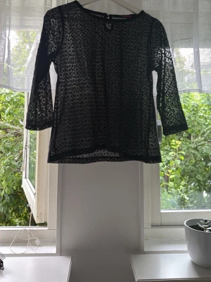 Svart spetsblus från Indiska - Säljer en svart spetsblus från Indiska med trekvartsärm och rund hals. Blusen är  i ett fint spetsmönster och har knapp i nacken. Perfekt att styla lager på lager eller med ett linne under.
