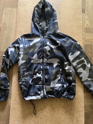 Klassisk camo vindjacka med huva - Snygg vindjacka i klassiskt camouflage-mönster med dragkedja framtill och huva. Jackan har elastiska muddar vid ärmarna och dragsko nedtill för extra passform. Perfekt för dig som gillar streetwear och vill ha en lätt jacka till vardagen.