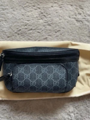 GG magväska - Snygg svart midjeväska från GG med klassiskt GG-monogram i grått. Väskan har dragkedja upptill, en mindre ficka framtill och detaljer i svart läder. Perfekt för att bära det viktigaste och ger en trendig touch till din outfit.