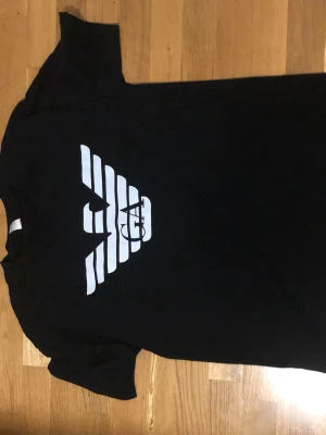 Armani T-shirt  - Säljer en svart t-shirt med en stor vit grafisk logga på bröstet. T-shirten har rund hals och korta ärmar. Materialet ser ut att vara bomull eller bomullsblandning, perfekt för en avslappnad och stilren look.