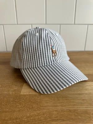 Polo Ralph Lauren Randig keps med broderad logga - Snygg keps med smala vita och gråa ränder samt en broderad polospelare på framsidan. Klassisk böjd skärm och justerbar rem bak. Tillverkad i ett lätt tyg som känns luftigt och fräscht, perfekt för en avslappnad stil. Kom gärna med bud