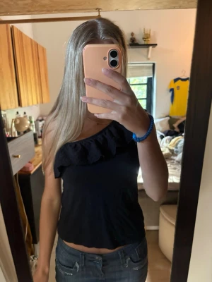 Svart oneshoulder topp med volang - Snygg svart oneshoulder topp med bred volangdetalj längs halsringningen. Toppen är ärmlös och har en axelbandslös sida. Tillverkad i mjukt bomullsmaterial som känns skönt mot huden. Perfekt att styla med jeans eller kjol.