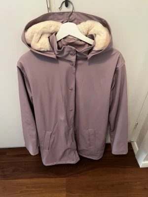 Ljuslila regnjacka med foder - Snygg ljuslila regnjacka från KappAhl med mjukt, beige teddyfoder i både huva och insida. Jackan har tryckknappar framtill, två fickor och en rymlig huva. Perfekt för regniga dagar när du vill hålla dig både torr och varm.