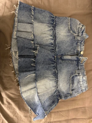 Blå jeanskjol med volang  - Säljer en kort blå jeanskjol med volang och råa kanter. Kjolen har klassiska fickor fram och bak, bälteshällor och dragkedja med knapp. Perfekt för dig som gillar en avslappnad och trendig stil.