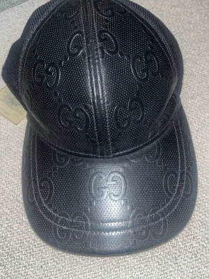 Svart keps i läder från Gucci - Snygg svart keps från Gucci i läder med det klassiska GG-mönstret över hela kepsen. Kepsen har böjd skärm och mesh på sidorna för extra ventilation. Perfekt för dig som vill ha en exklusiv och trendig accessoar.