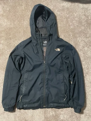 Svart vindjacka från The North Face - Snygg svart vindjacka från The North Face med huva och dragkedja framtill. Jackan har två fickor med dragkedja och elastiska muddar vid ärmsluten. Tillverkad i lätt och tåligt material, perfekt för blåsiga dagar.