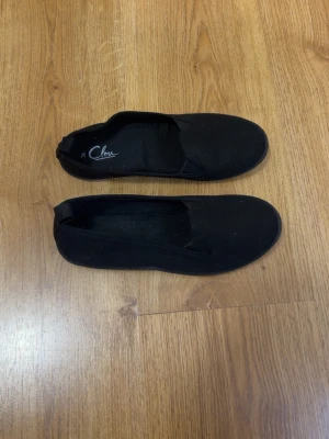 Svarta ballerinaskor från Clou - Säljer ett par klassiska svarta ballerinaskor från Clou. Skorna har en enkel och stilren design med rund tå och elastiska band över vristen för bättre passform. Tillverkade i textilmaterial med en mjuk sula som gör dem bekväma att använda hela dagen.