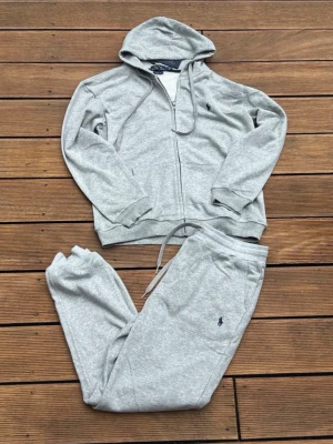  Ralph Lauren Tracksuit  - Mysigt grått loungewear-set från Polo Ralph Lauren med hoodie och matchande byxor. Setet har dragkedja, fickor och justerbar midja på byxorna. Båda delarna har den klassiska Polo-loggan broderad. Perfekt för chill dagar hemma.