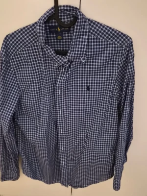 Blårutig skjorta från Ralph Lauren - Klassisk blårutig skjorta från Ralph Lauren med button-down krage och broderad logga på bröstet. Skjortan har långa ärmar och är tillverkad i mjuk bomull. Perfekt för en stilren och avslappnad look.