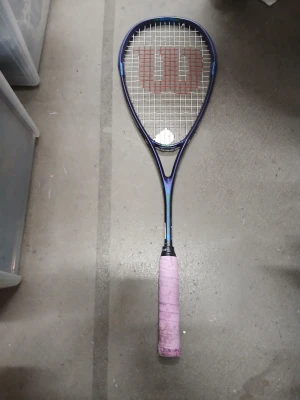 Wilson squashracket / tennisracket - Säljer ett squashracket från Wilson. Racketen har en lila färg med gröna och blå detaljer. Greppet är lindat med ett rosa greppband, rekommenderar att greppa om. Strängarna är vita med röda strängar i mitten som bildar en "W"-logotyp.