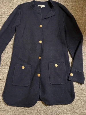 Mörkblå cardigan från Hampton Republic - Mörkblå lång cardiganjacka från Hampton Republic med guldknappar framtill och på fickorna. Två stora fickor fram och rundad nederkant. Stickad i mjukt material och har lång ärm. Enkel och stilren design som passar till mycket.