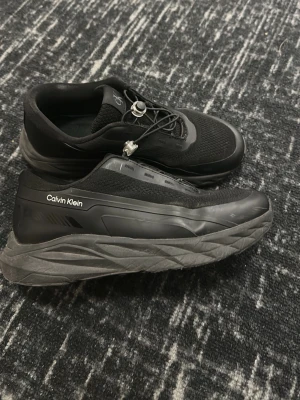 Svarta sneakers från Calvin Klein - Stilrena svarta sneakers från Calvin Klein med chunky sula och meshdetaljer. Skorna har elastiska snören med snabbspänne och diskret CK-logga på plösen samt Calvin Klein-tryck på sidan. Materialmix av textil och syntet ger en modern look.