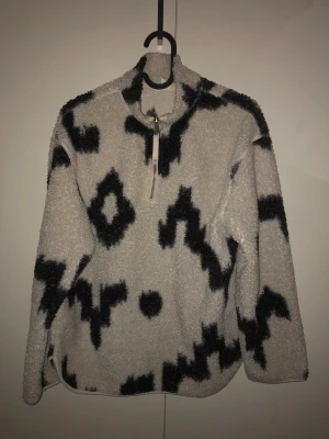 Beige och svart mönstrad fleece - Säljer en beige stickad fleece med svart grafiskt mönster. Tröjan har halv dragkedja framtill, hög krage och är gjord i mjukt, fluffigt material. Perfekt för dig som gillar en avslappnad och trendig stil.