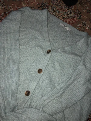 Ljusblå kofta från NA-KD med knappar - Mysig ljusblå kofta från NA-KD med v-ringning och tre bruna knappar framtill. Koftan är stickad i ett mjukt material och har en avslappnad passform. Perfekt att slänga på sig när det är lite kyligt ute.