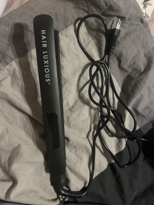 Svart plattång Hair Luxious - Säljer en svart plattång från Hair Luxious med sladd och digital display. Plattången har knappar för temperaturreglering och är tillverkad i plast och metall. Perfekt för att styla håret snabbt och enkelt. Man får med sig kartongen.