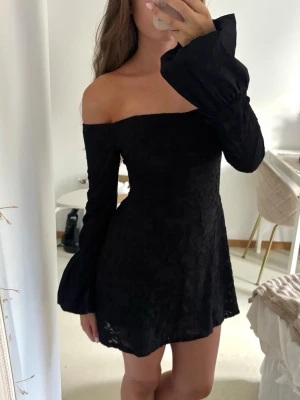 Svart offshoulder kortklänning - Snygg svart kortklänning med offshoulder-design och långa ärmar. Klänningen har en lätt utsvängd kjol och diskret blommönstrad struktur i tyget. Perfekt för dig som vill ha en elegant och trendig look. Ny med prislappen kvar. 