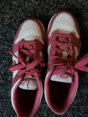 Nike Dunk Low rosa och vita sneakers - Snygga Nike Dunk Low sneakers i rosa och vitt med klassisk swoosh på sidan. Skorna har perforerad tåbox, platta rosa skosnören och lågt skaft. Ovandelen är i läder och insidan är fodrad i matchande rosa tyg. Perfekta för dig som gillar streetwear och sportig stil.